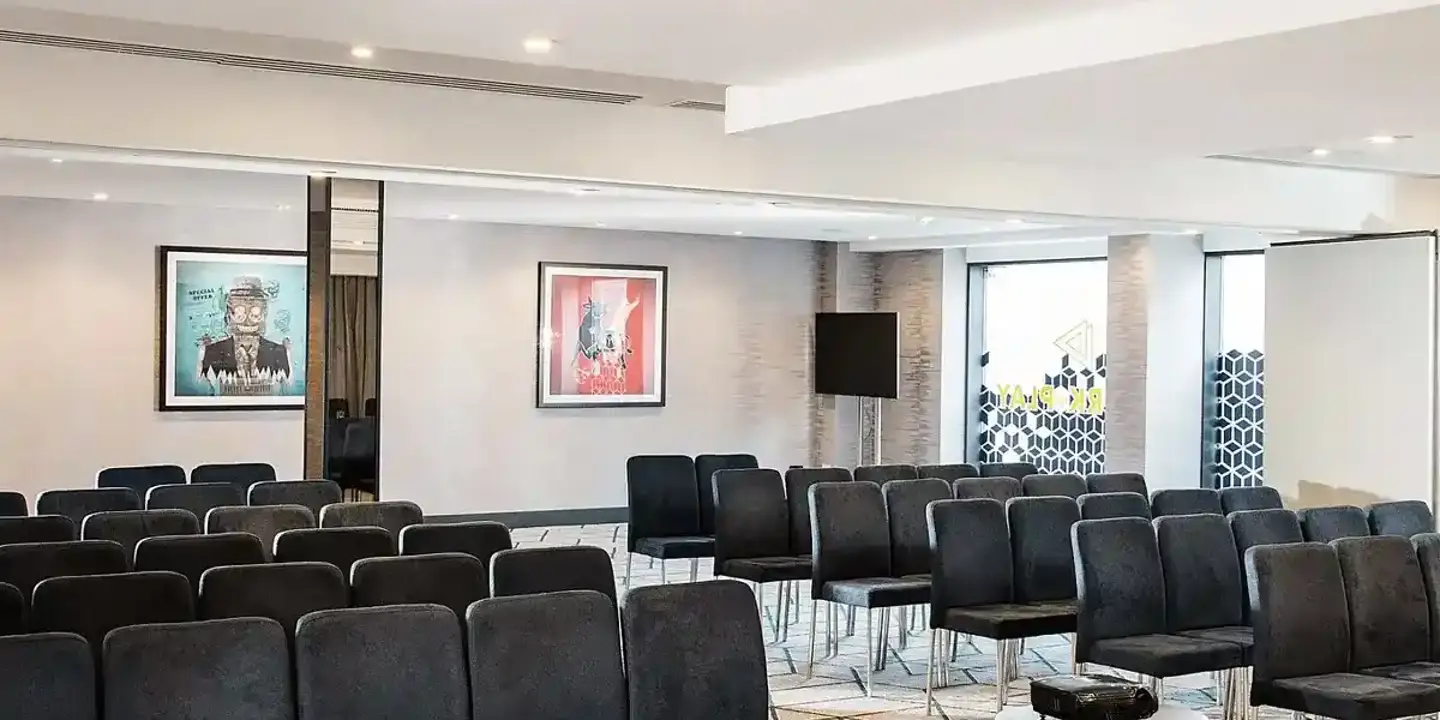 A meeting room set up for a seminar with rows of black fabric and metal seats, a geometric patterned cream and grey carpet, and a projector.