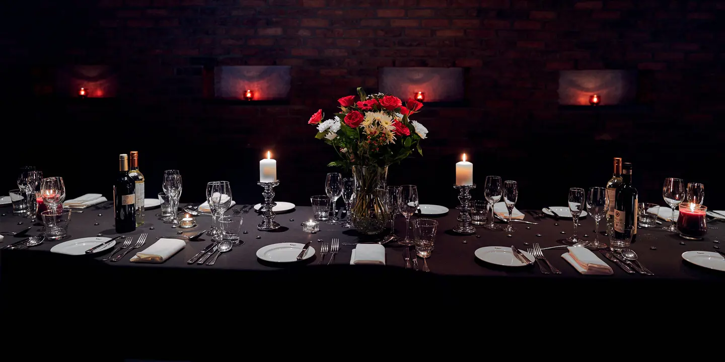 Wide angle view of Private dining room featuring a table laid for dinner service, white candles in silver candlesticks, and a red and white floral centrepiece. The back wall is exposed brick with 4 lit candles in cubby holes.