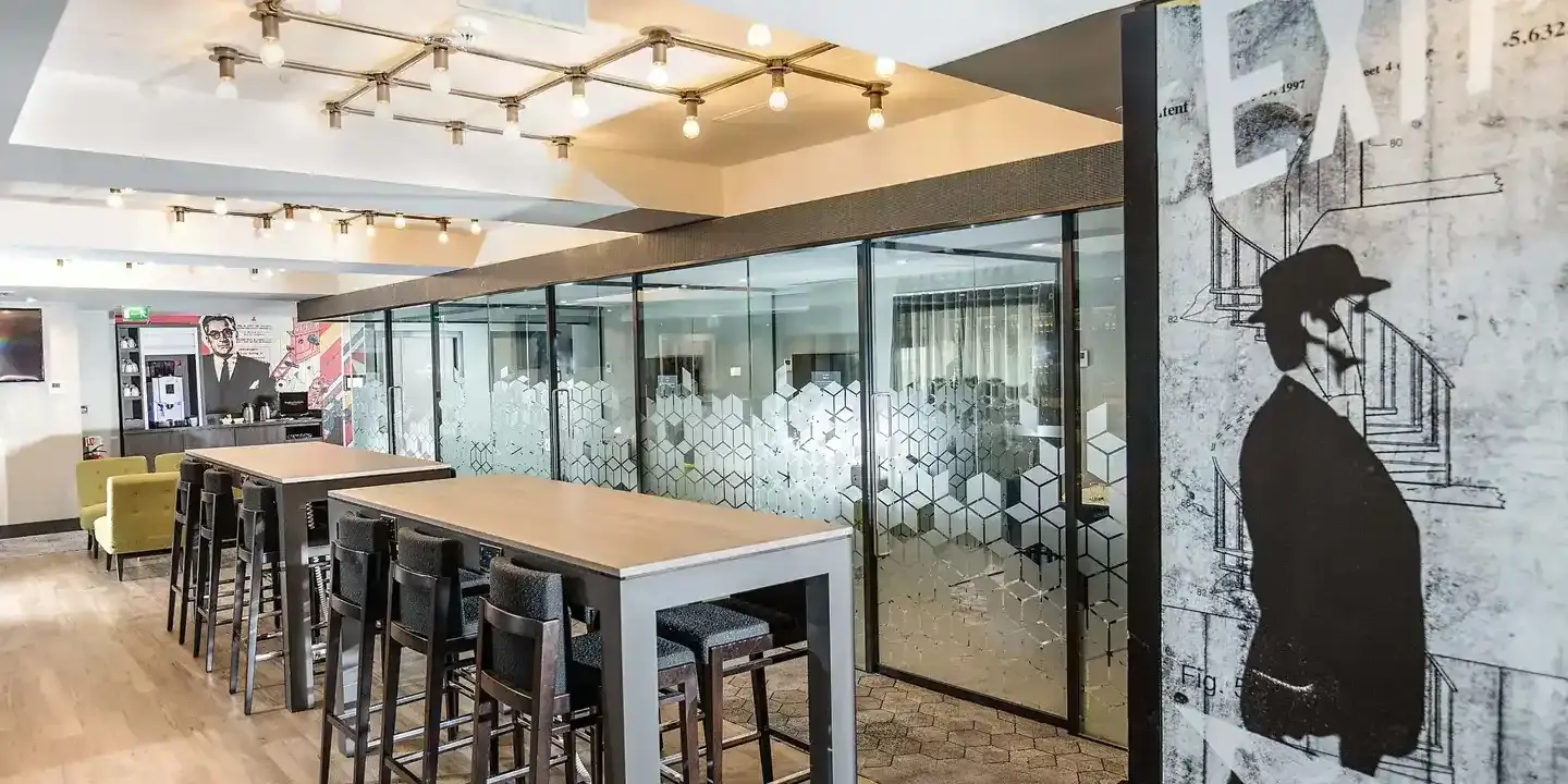 Malmaison Birmingham Work Plus Play Meeting Room  with stencil graffiti adorning a wall, tables with stools, and glass partitioned meeting areas. 