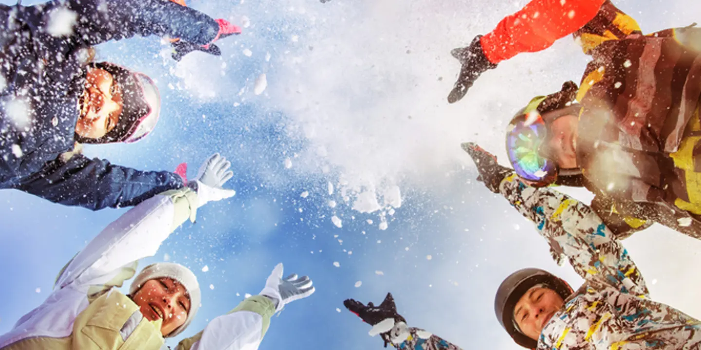 A photo from below of a circle of people in ski wear throwing snow into the air whilst looking down at the camera 
