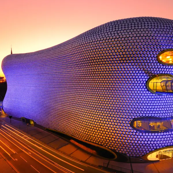 The Birmingham bull ring at dusk lit up in blue and purple with traffic whizzing by