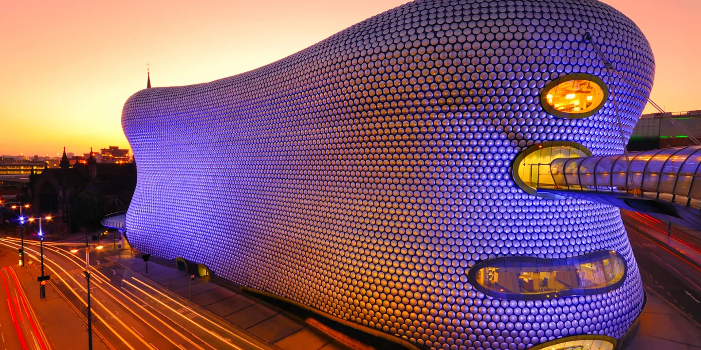 The Birmingham bull ring at dusk lit up in blue and purple with traffic whizzing by
