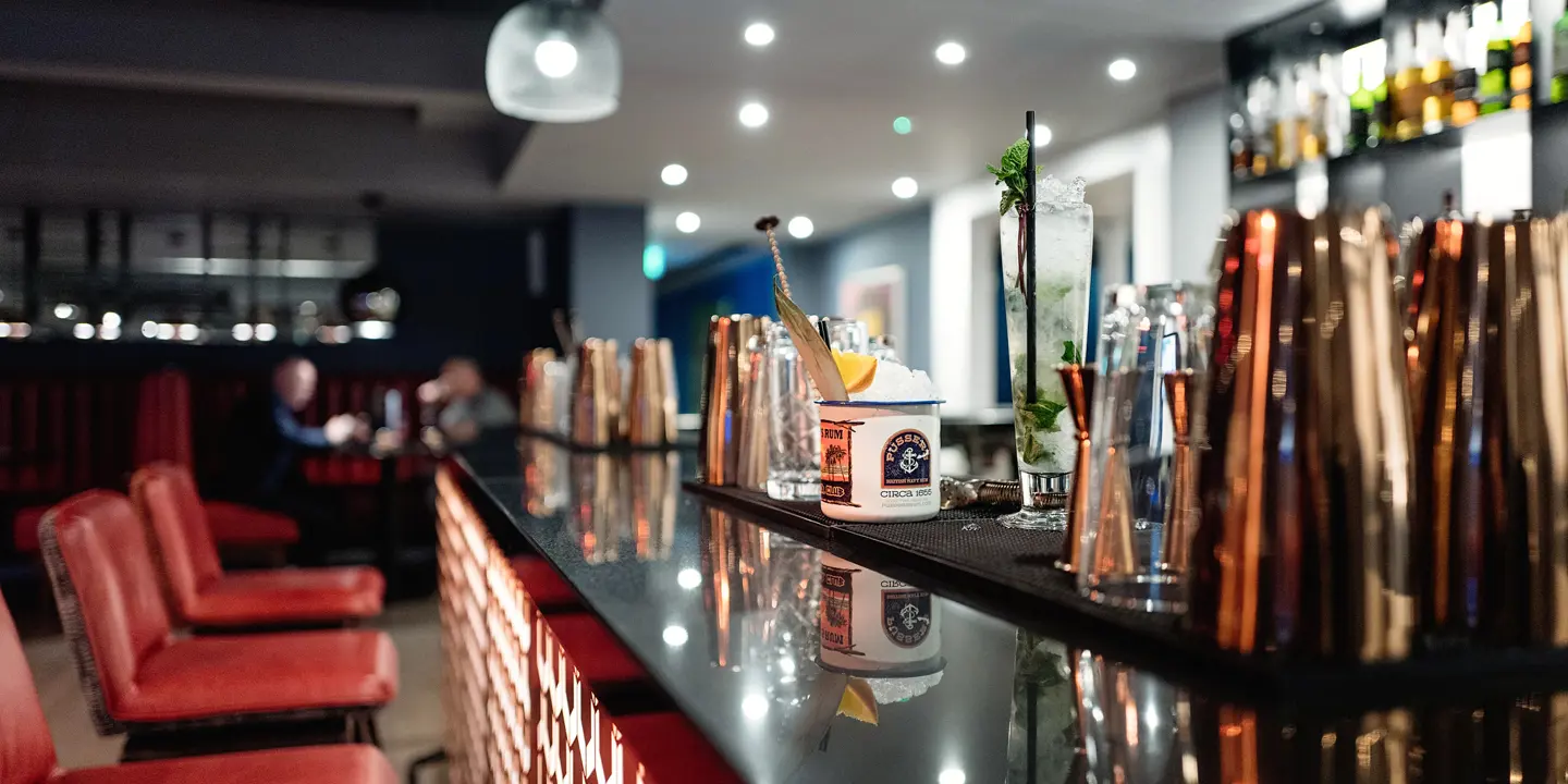 A tall cocktail displayed on a glossy black bar counter, with red leather stools, and brass tumblers out of focus.