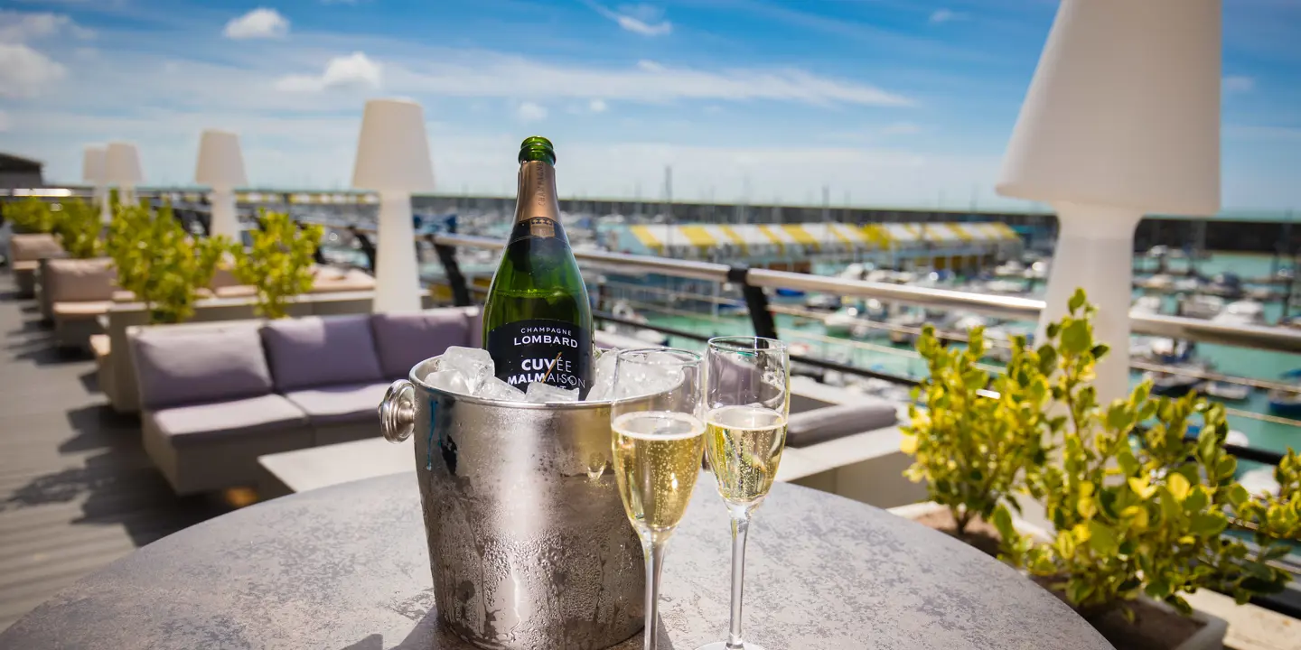 A bottle of champagne and two wine glasses on a table on a wooden deck terrace, with Brighton Marina in the background.