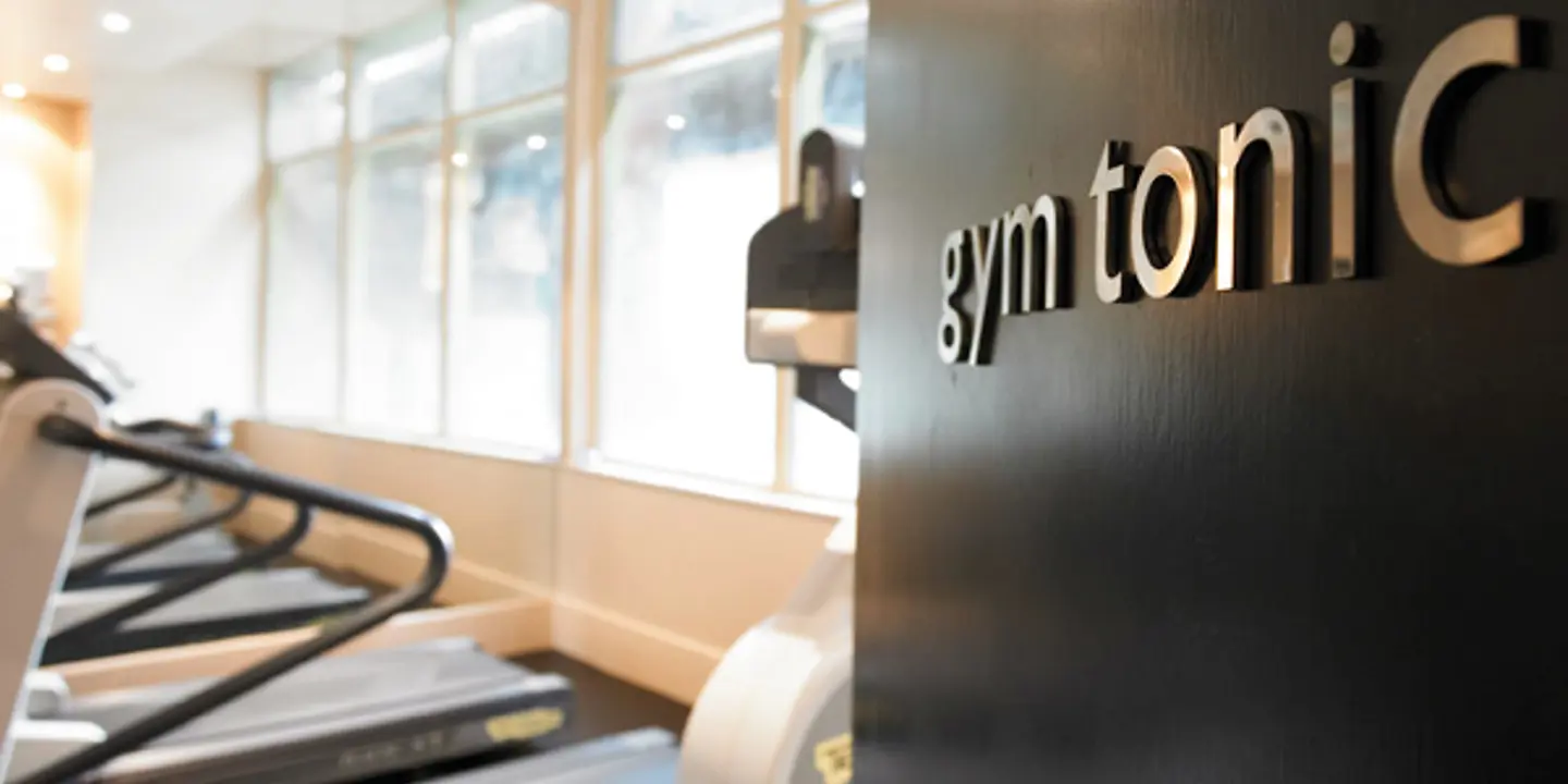 Black door featuring silver wording reading "gym tonic". The door is ajar showing part of a room featuring gym equipment including a row of treadmills