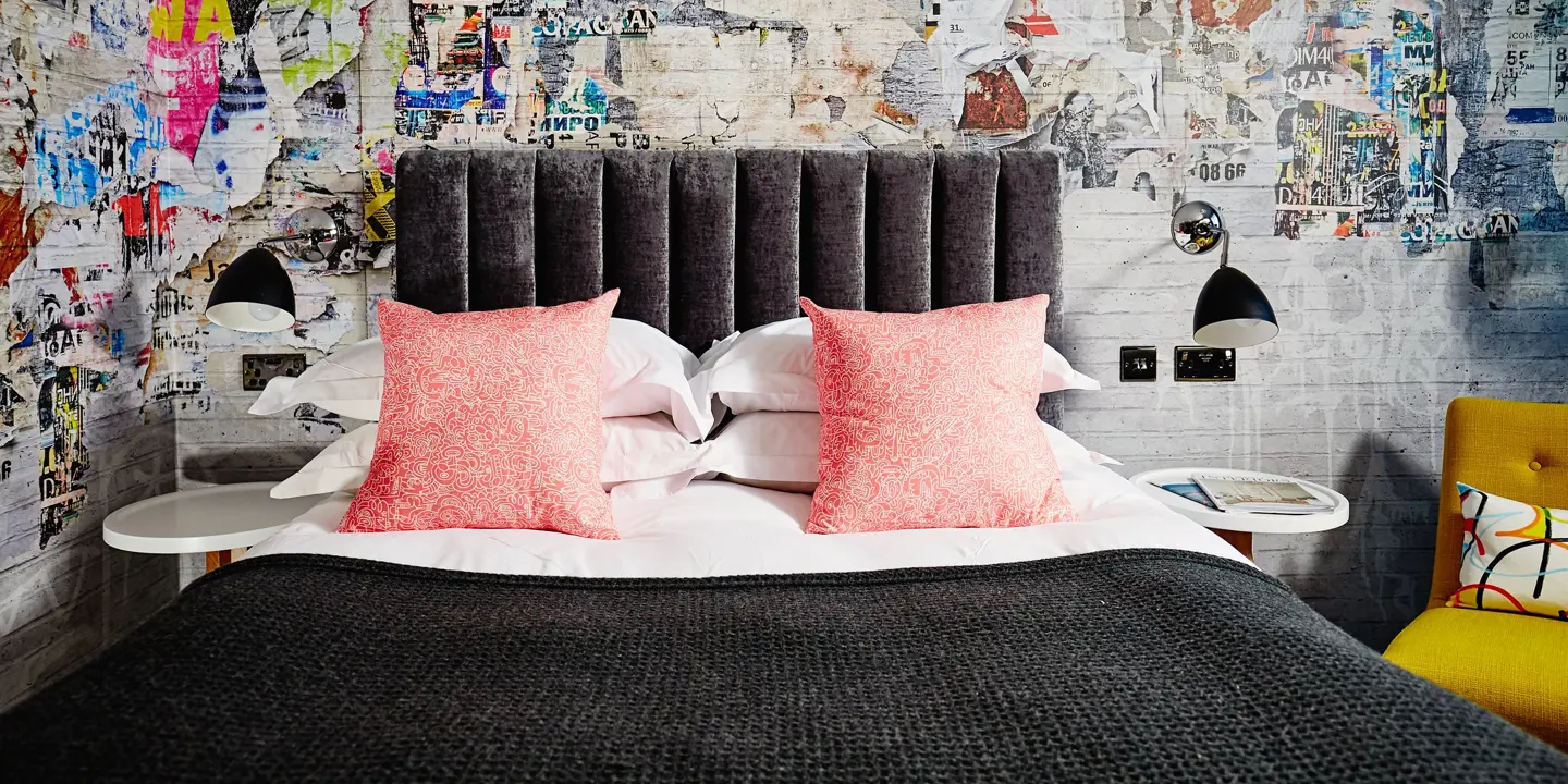 Grey bed with 2 pink pillows and a brightly decorated wall. 