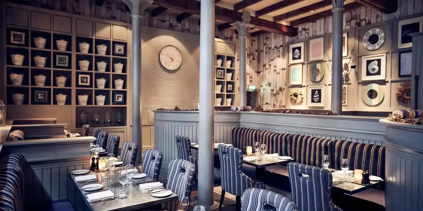 Alternate angle of Malmaison Belfast restaurant interior showing grey feature pillars, blue cushioned bench seating, wooden tables, and a wall adorned with a clock, shelving units and artwork.