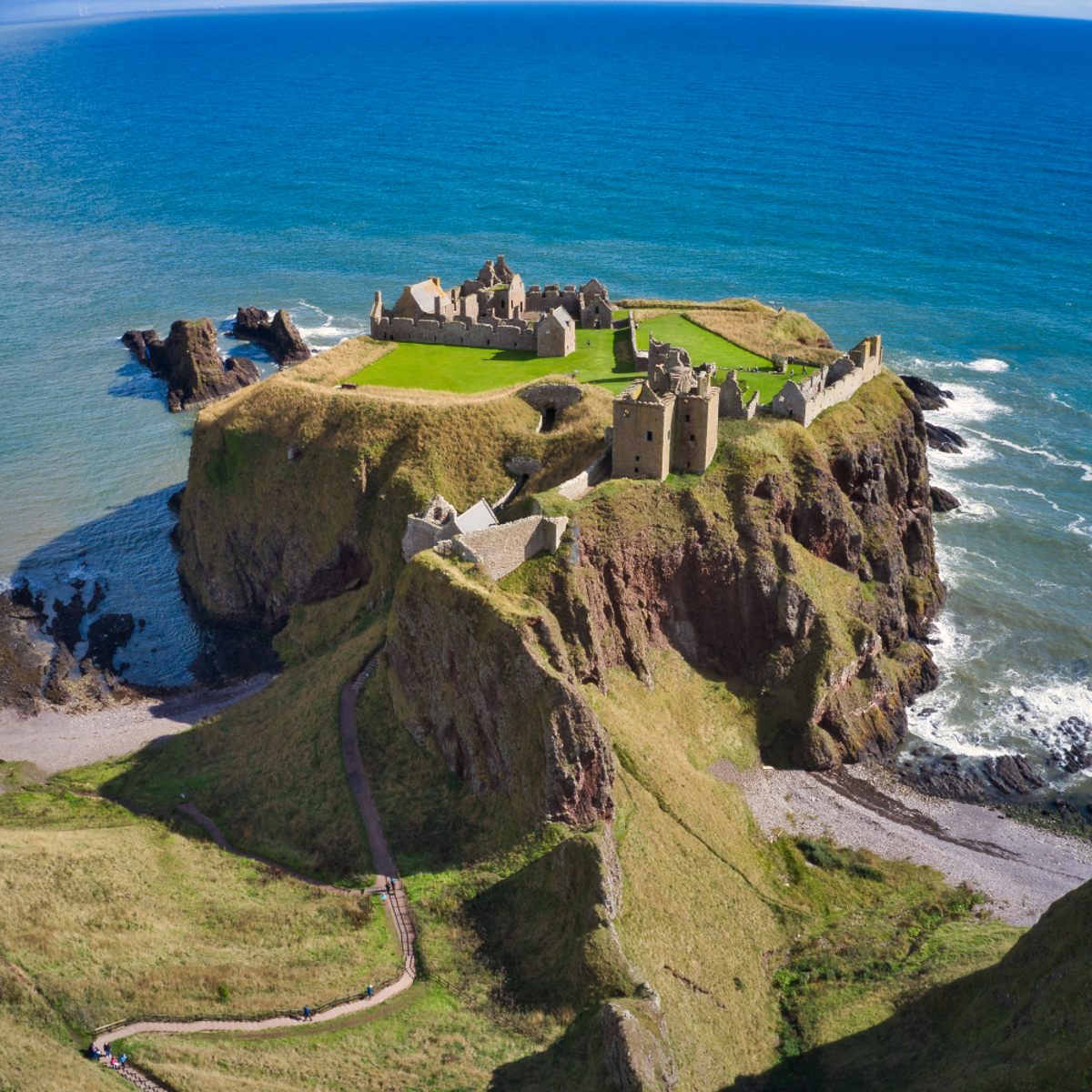 An aerial shot giving a birds-eye view of Dunnottar Castle on a sunny day. The winding cliff path is prominent making its way through the grassy meadows. The sea is vibrantly coloured with several shades of blue.