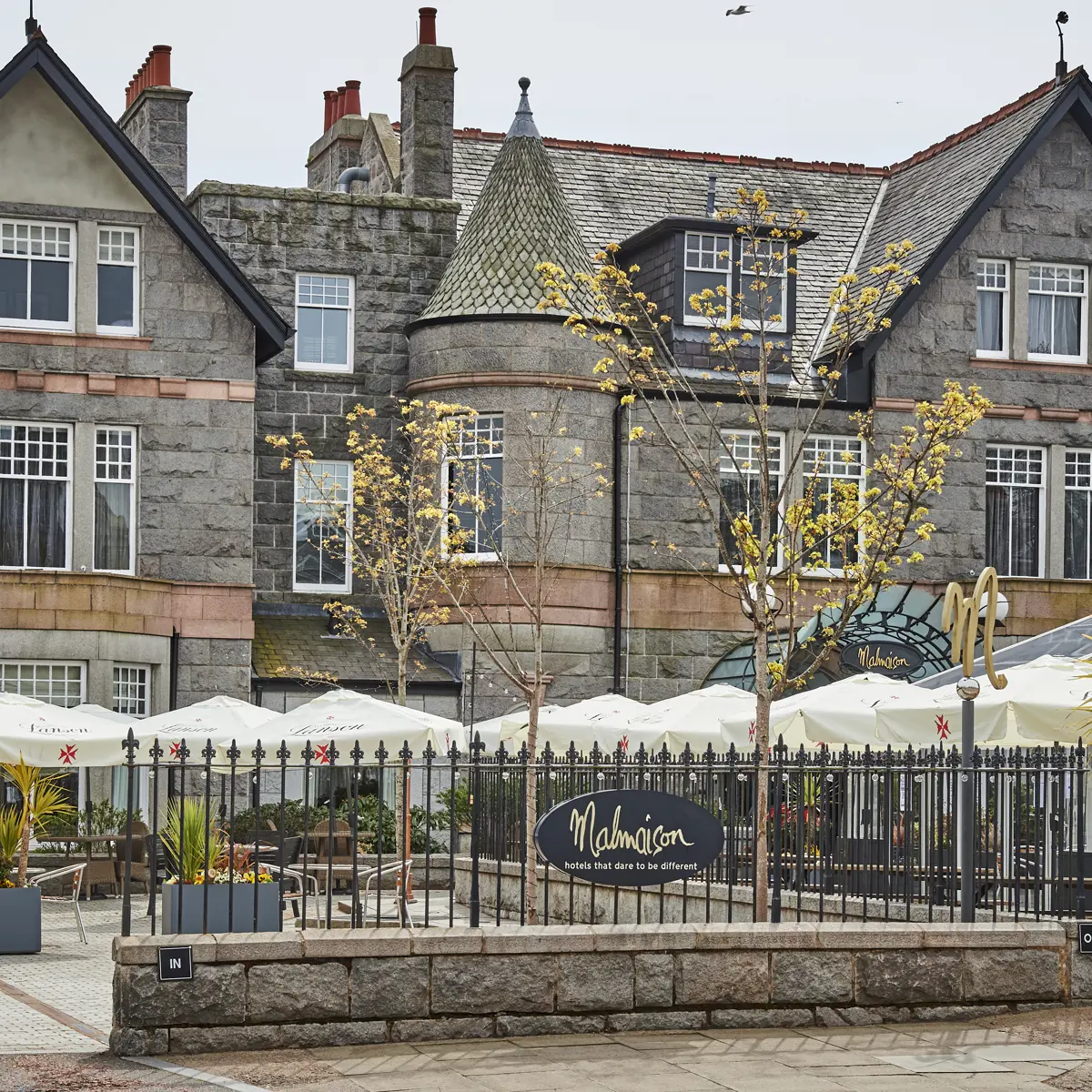 Al fresco dining area outside of Malmaison Aberdeen, featuring plentiful wooden and metal seating and tables, multiple white umbrellas, and a conservatory area behind ornate black metal railing
