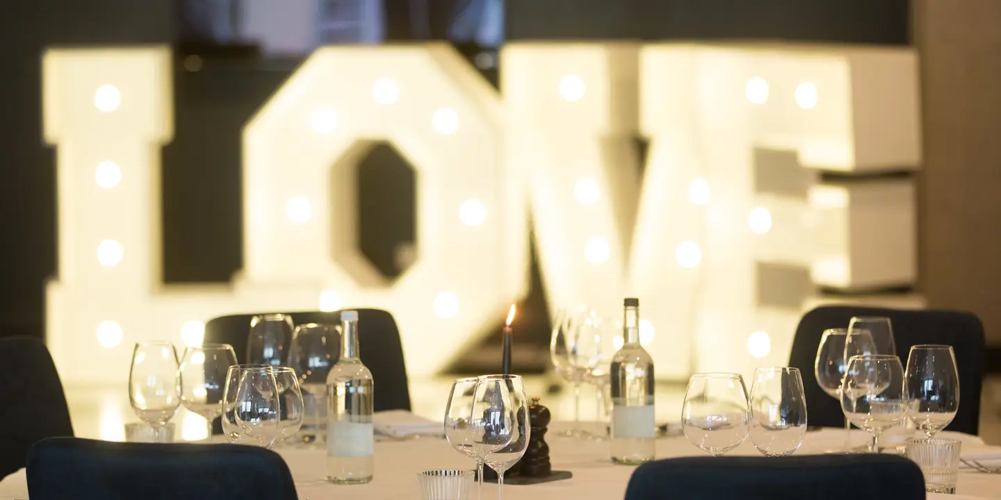A table arranged with wine glasses and place settings. A light display in the background spells out the word love