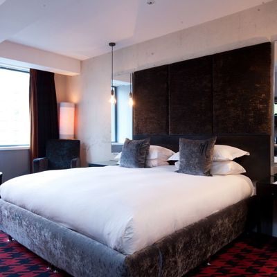 View of the bed in the standard Malmaison Birmingham hotel. White linen sheets, grey throw cushions, and brown velvet head rest.