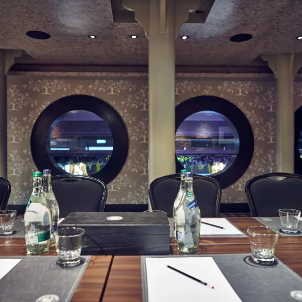 Luxurious meeting room showing a close up of a wooden table adorned with bottles of water and glasses, with 5 black chairs in focus, and 3 black framed porthole style windows in the background.