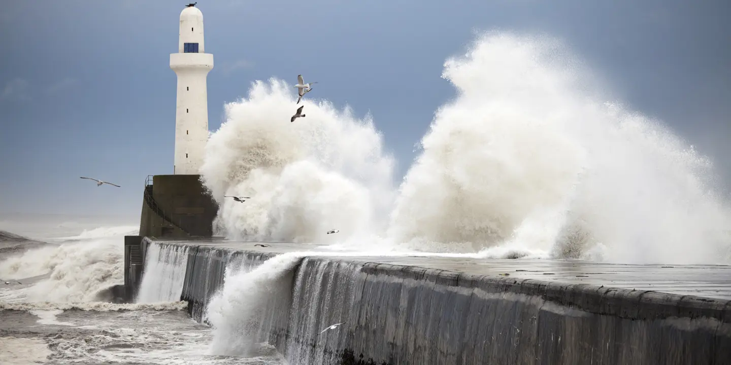 A dramatic image of white sea waves crashing over a grey stone pier, a white lighthouse is visible at the end of the pier and seagulls wheel across the sky.