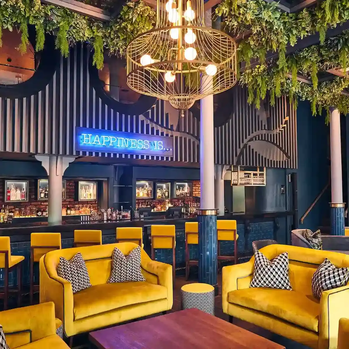 Yellow fabric sofas and bar stools prominently feature around a wooden table, with the bar in the background and a blue neon sign reading "happiness is" is displayed above the bar.