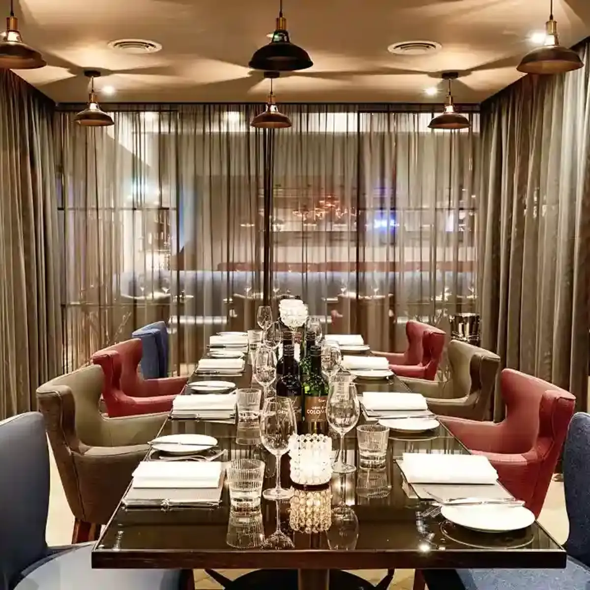 Another angle of The Cage dining room featuring a lengthy table adorned with blue, red and gold chairs, curtained glass walls, and brass light fixtures.