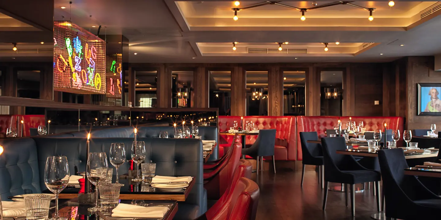 Birmingham Malmaison restaurant interior alternate view showing red leather seating including booths. A feature neon sign with multiple colours is visible on one wall. 