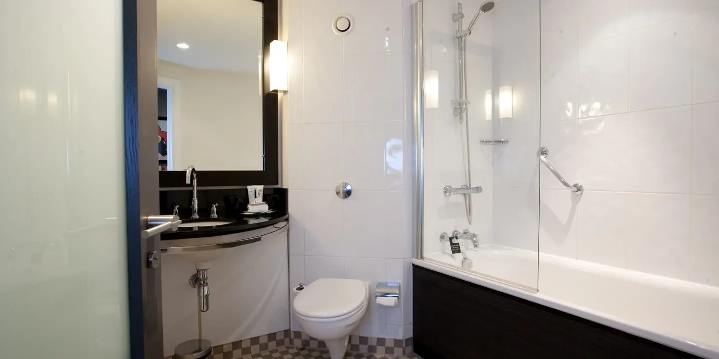 A bathroom featuring a white bathtub with built in silver shower and glass screen. A white toilet and black sink are visible.